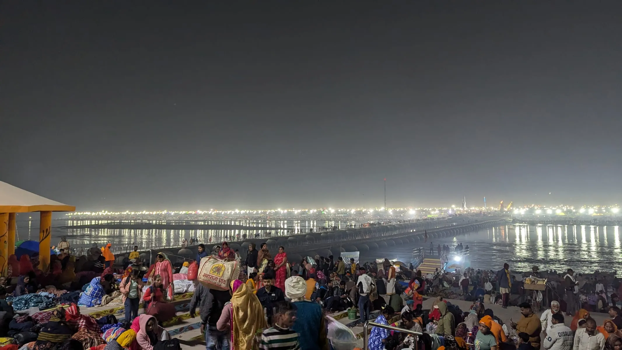 Crowds of pilgrims on the riverbank at night, with the lights of the Kumbh camps stretching along the water